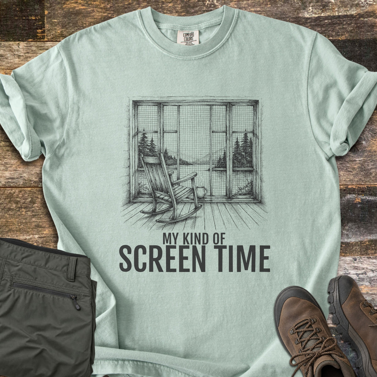 My Kind Of Screen Time Comfort Colors T-shirt in Bay, laid flat. A dark gray sketch of a rocking chair and steaming mug before a screened window looking out on a lake and pines, above the words My Kind Of Screen Time. Olive hiking pants and brown hiking boots rest on weathered wood planks.