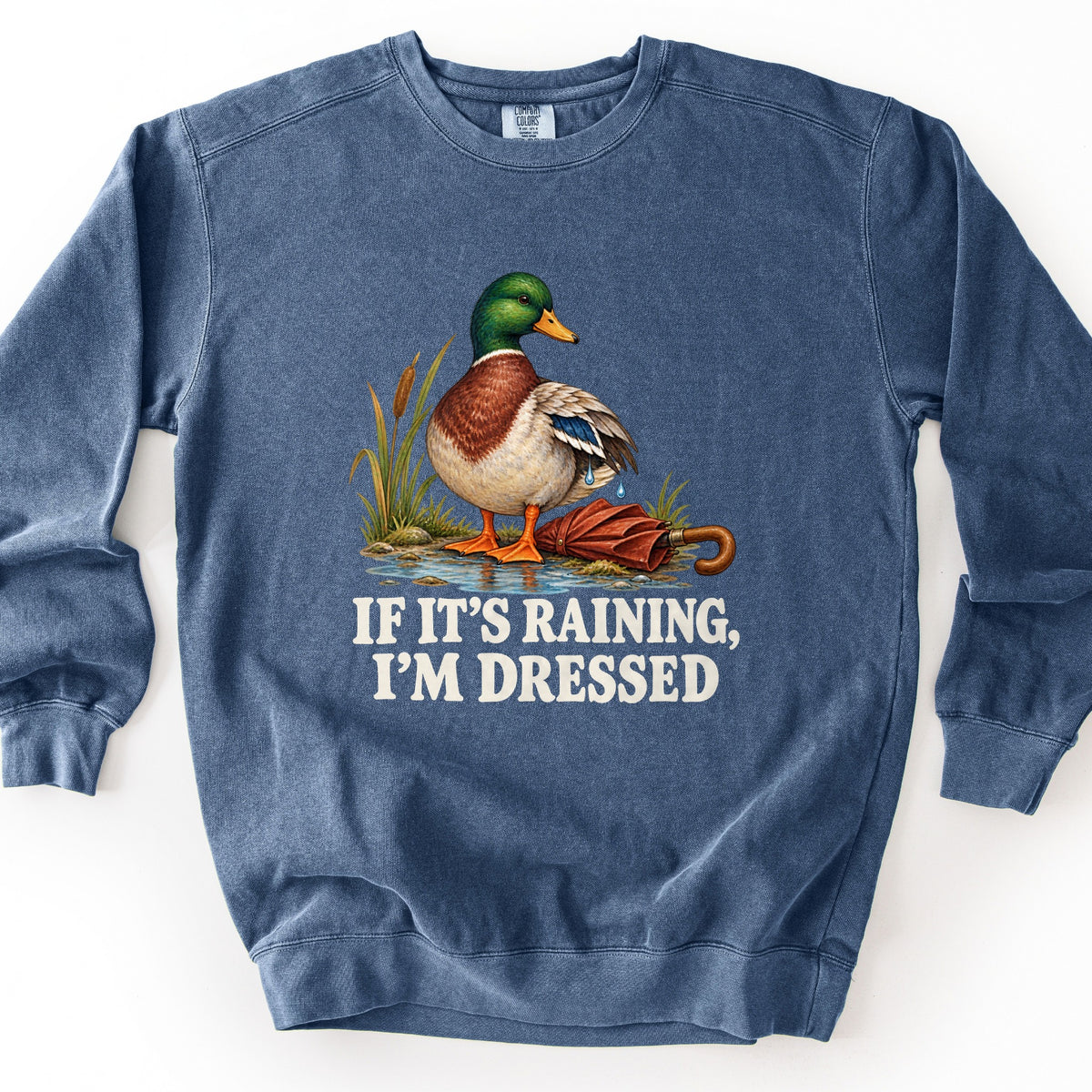 If It's Raining, I'm Dressed Comfort Colors crewneck sweatshirt in Denim, laid flat. An illustrated mallard duck stands at a puddle's edge among cattails beside a closed red umbrella, water dripping from its wing, above the words If It's Raining, I'm Dressed. Laid on a white background.