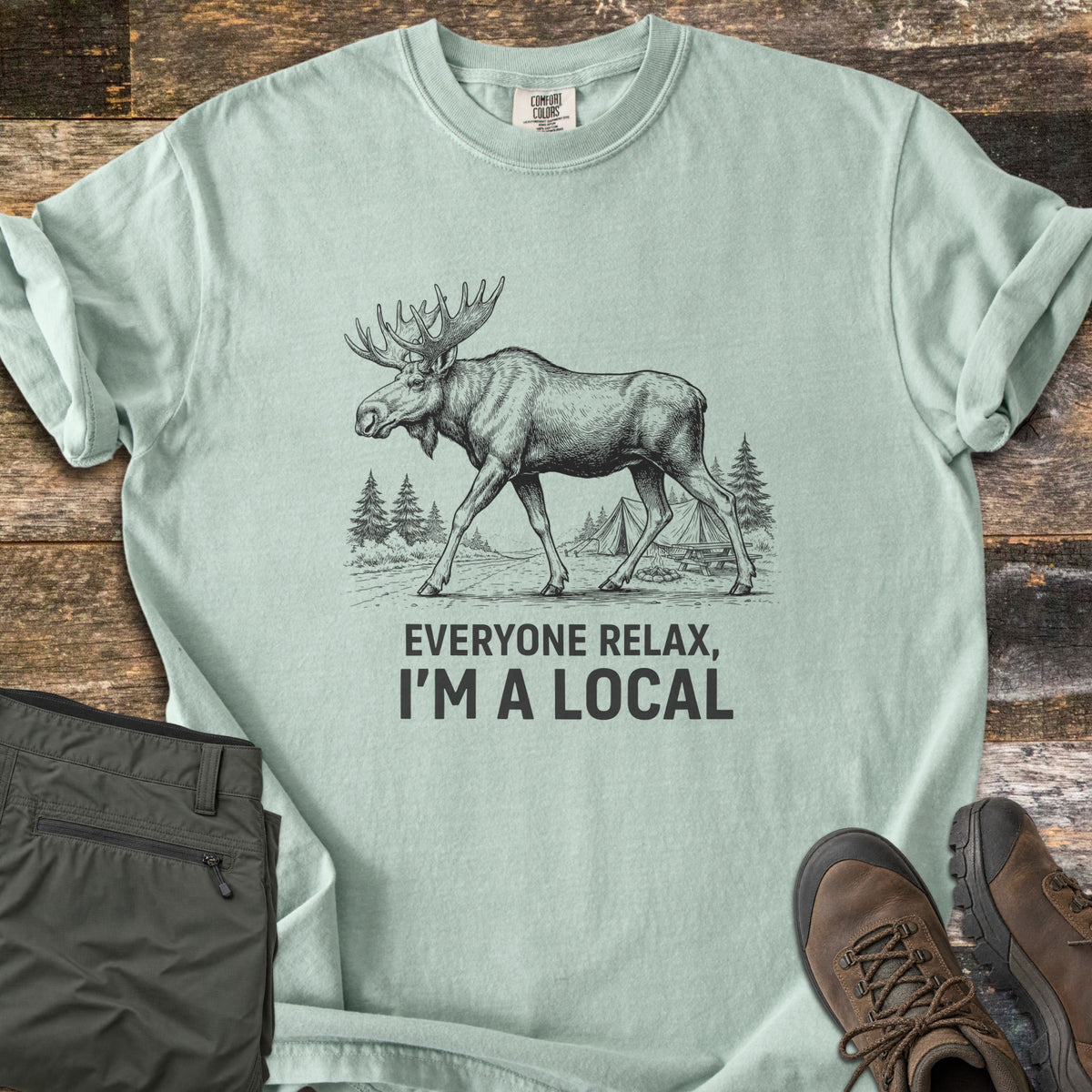 Everyone Relax I'm A Local Comfort Colors T-shirt in Bay, laid flat. A sketched moose walks past pine trees and tents at a campsite above the words Everyone Relax, I'm A Local. Sleeves are rolled on a wood floor beside folded green pants and brown hiking boots.