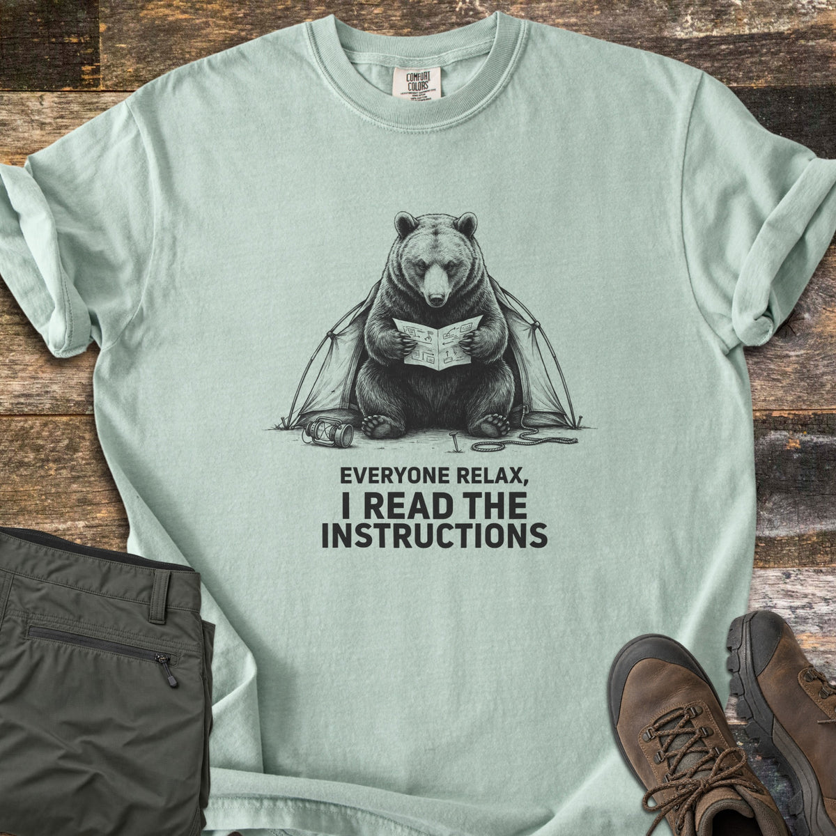 Everyone Relax I Read The Instructions Comfort Colors T-shirt in Bay, laid flat. A black sketch of a bear reading instructions in front of a tent, with a tipped lantern and rope, above the words Everyone relax, I read the instructions. Olive cargo pants and brown hiking boots sit on dark wood.