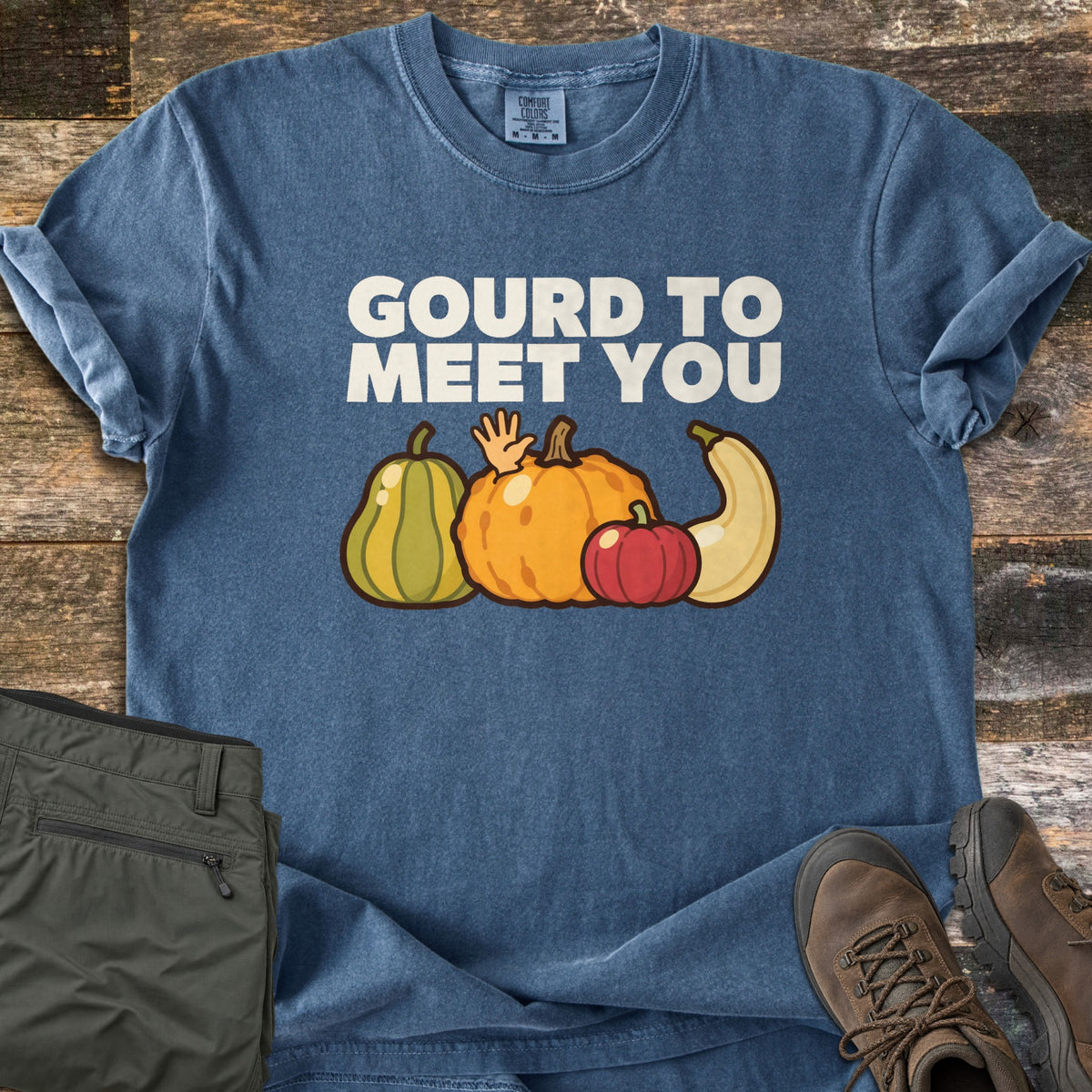 Gourd To Meet You Comfort Colors T-shirt in Denim, laid flat. White text reads Gourd To Meet You above four cartoon gourds, with a hand waving from behind an orange pumpkin. Olive cargo shorts and brown hiking boots lie beside it on weathered wood planks.