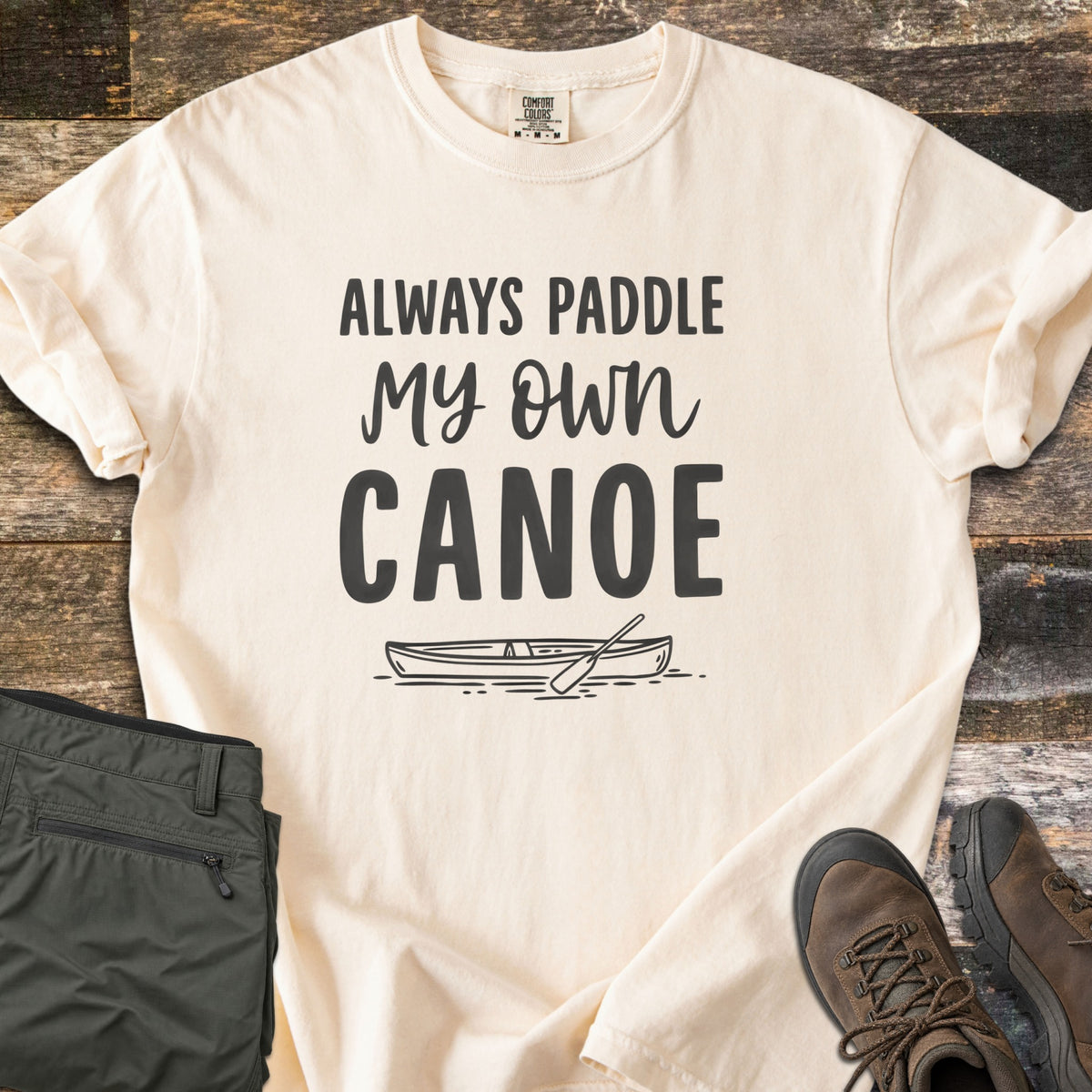 Paddle My Own Canoe Comfort Colors T-shirt in Ivory, laid flat. Charcoal text reads Always Paddle My Own Canoe above a line drawing of a canoe with a paddle on the water. It lies on weathered wood with rolled sleeves, folded olive hiking pants, and brown hiking boots.