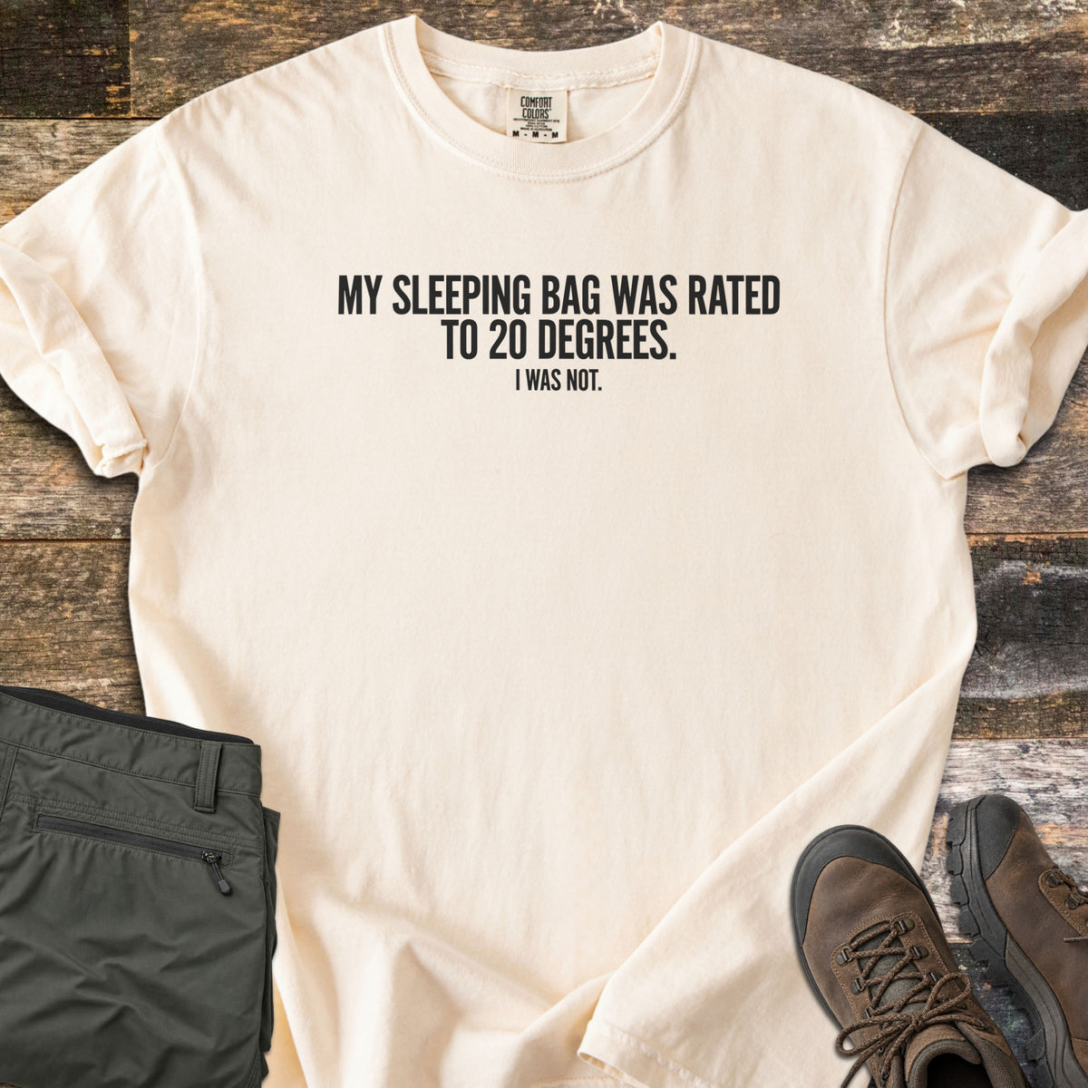 Rated To 20 Degrees Comfort Colors T-shirt in Ivory, laid flat. Black text across the chest reads My Sleeping Bag Was Rated To 20 Degrees. I Was Not. The shirt has rolled sleeves and lies on weathered wood planks beside folded olive hiking pants and brown hiking boots.