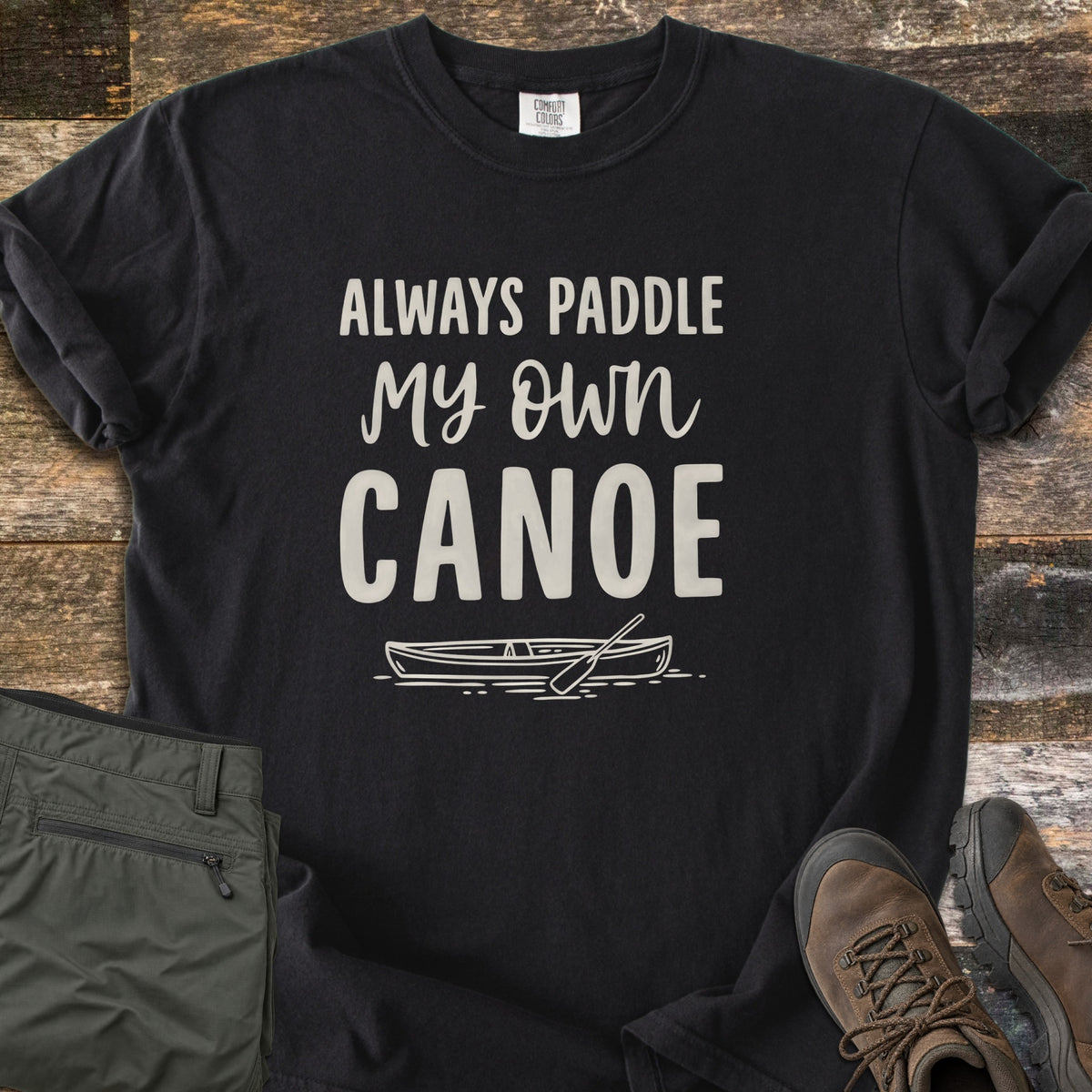 Paddle My Own Canoe Comfort Colors T-shirt in Black, laid flat. Cream text reads Always Paddle My Own Canoe above a line drawing of a canoe with a paddle on the water. It lies on weathered wood with rolled sleeves, folded olive hiking pants, and brown hiking boots.