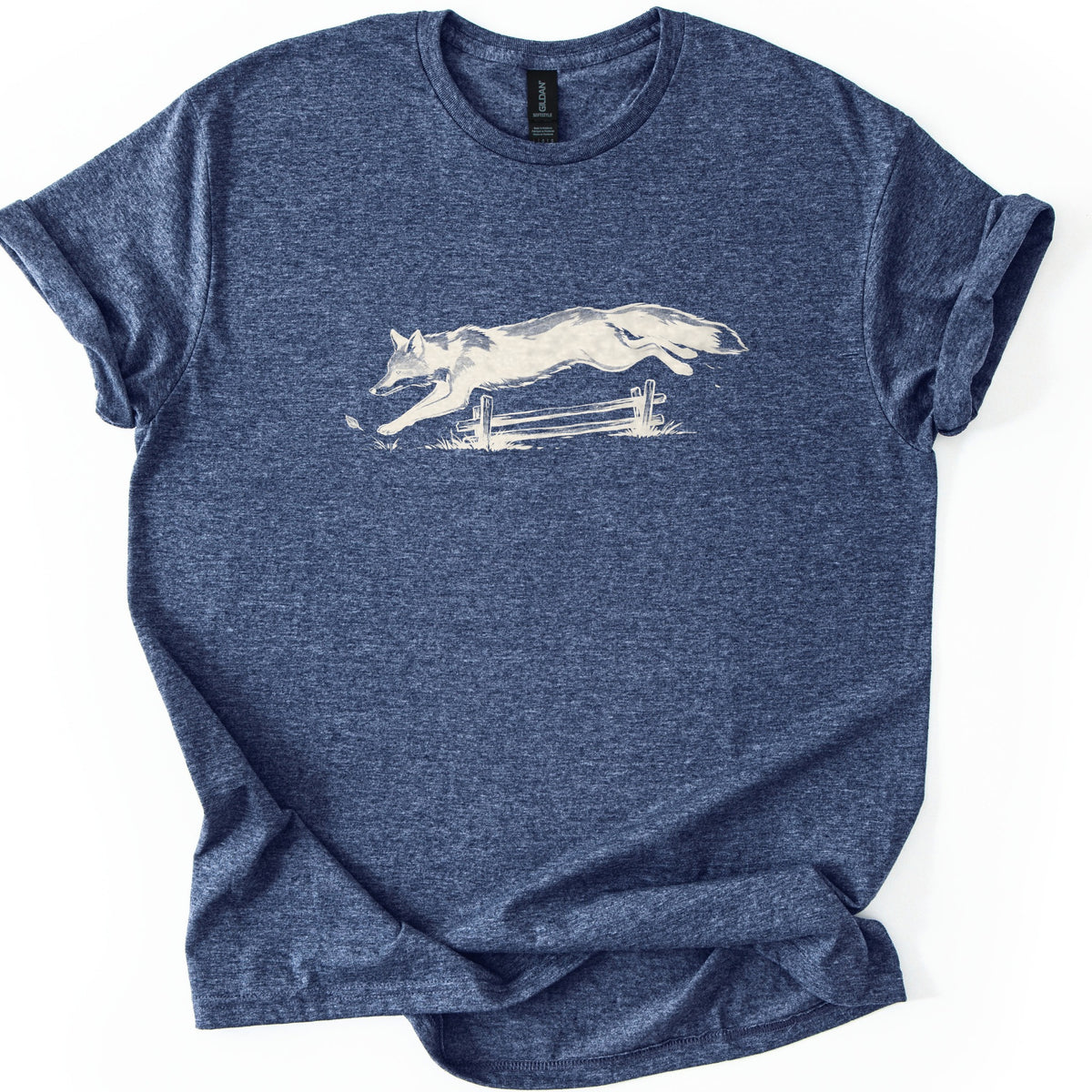 Leaping Fox Comfort Colors T-shirt in Heather Navy, laid flat. A cream-colored sketch of a fox leaping over a low wooden rail fence, with grass tufts and a falling leaf below. The shirt has cuffed sleeves and lies on a white background.