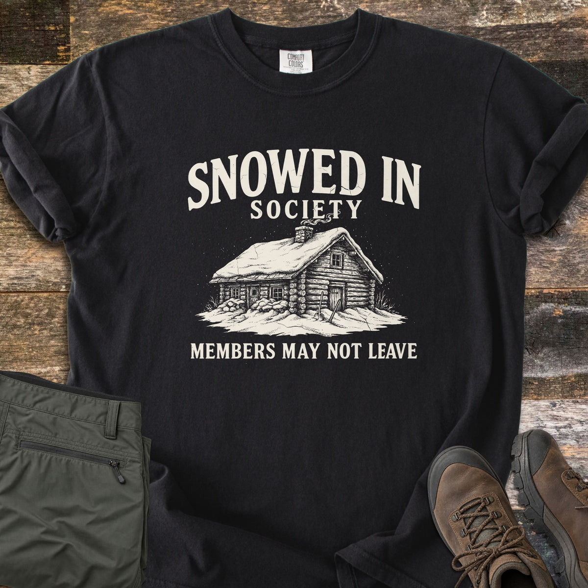 Snowed In Society Comfort Colors T-shirt in Black, laid flat. A cream print of a snow-covered log cabin with a smoking chimney sits under the words Snowed In Society and above Members May Not Leave. Folded olive pants and brown hiking boots lie beside it on weathered wood.