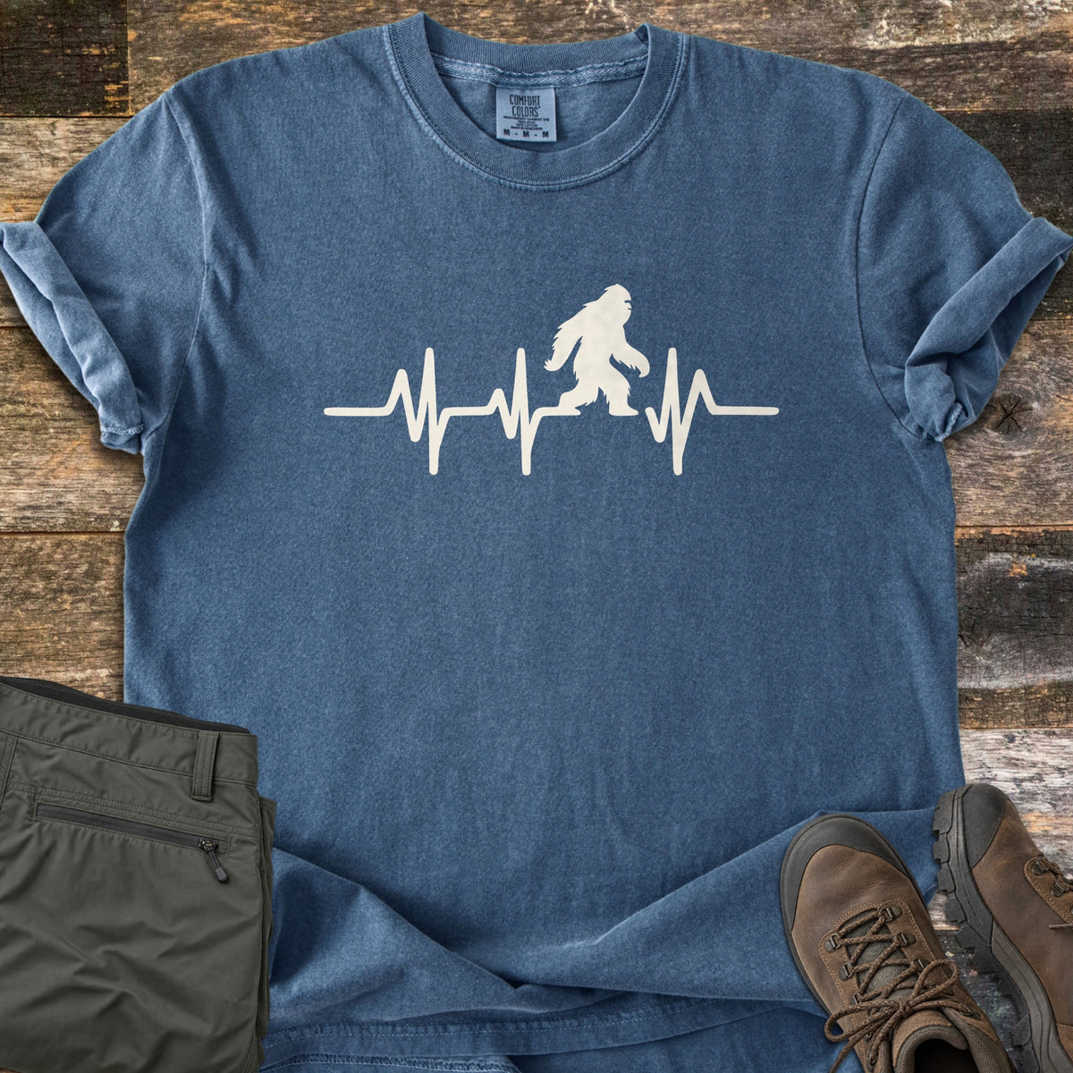 Bigfoot Heartbeat Comfort Colors T-shirt in Denim, laid flat. A cream heartbeat line runs across the chest with a walking Bigfoot silhouette in the middle; there are no words. The shirt lies on weathered wood beside folded olive hiking pants and brown hiking boots.