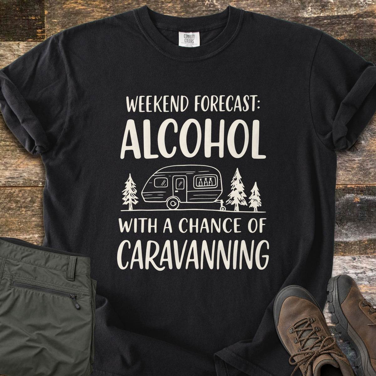 Alcohol With a Chance of Caravanning Comfort Colors T-shirt in Black, laid flat. Cream print reads Weekend Forecast: Alcohol above a caravan with bottles in its window between pine trees, then With a Chance of Caravanning. Olive trousers and brown hiking boots sit on dark wood planks.