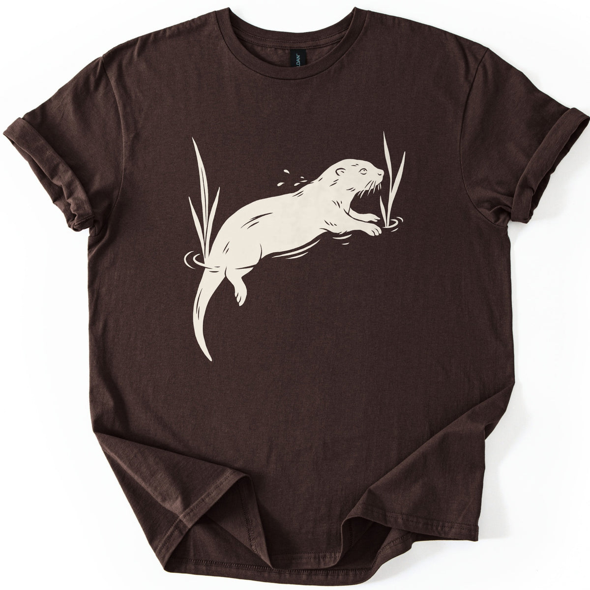 Swimming Otter Comfort Colors T-shirt in Dark Chocolate, laid flat. A cream linocut-style otter swims through rippling water between tall reeds, with droplets above its head. No words are printed. The sleeves are cuffed and the shirt lies on a white background.