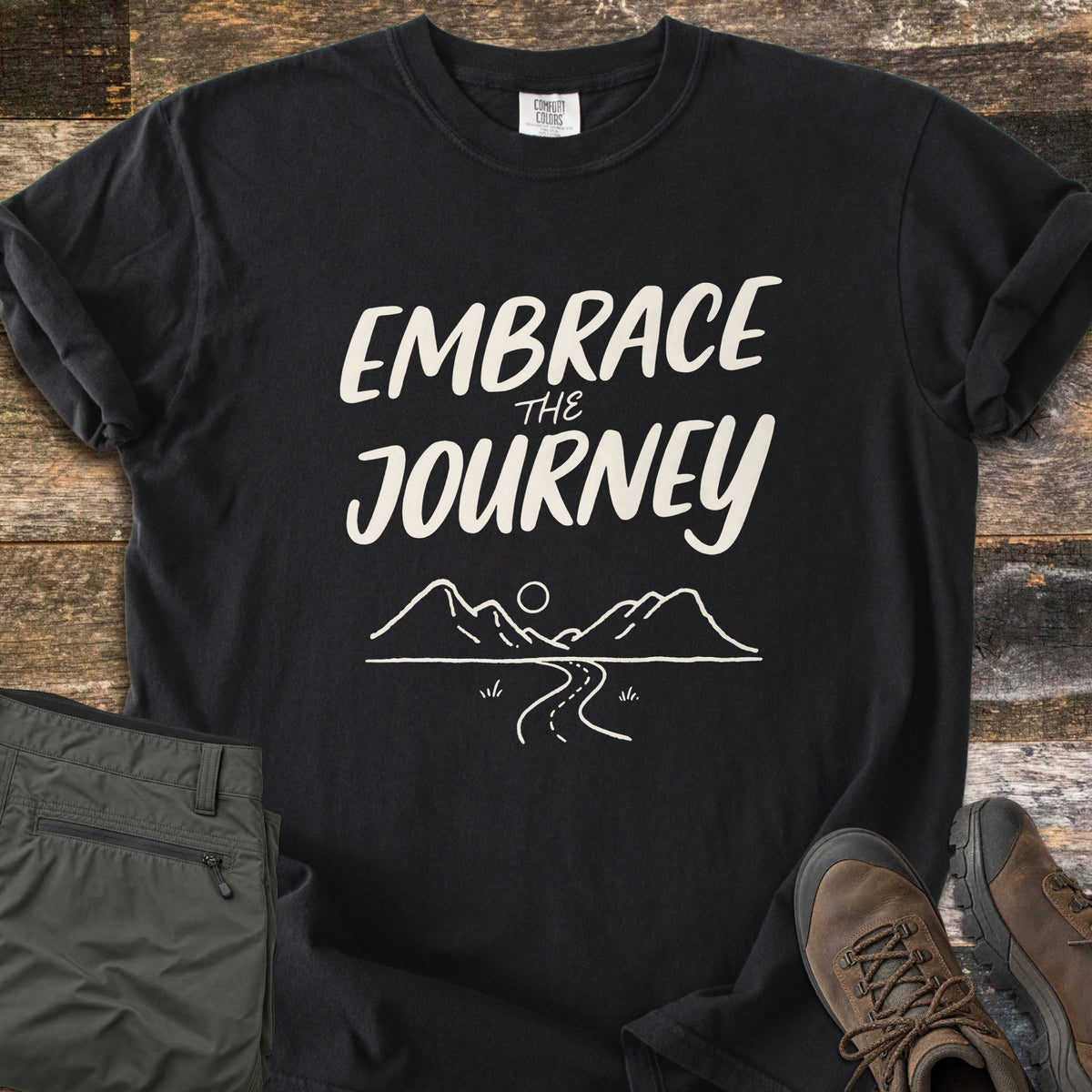 Embrace the Journey Comfort Colors T-shirt in Black, laid flat. Cream brush lettering reads Embrace the Journey above a line drawing of mountains, a sun, and a winding road. Sleeves cuffed on weathered wood beside folded green pants and brown hiking boots.