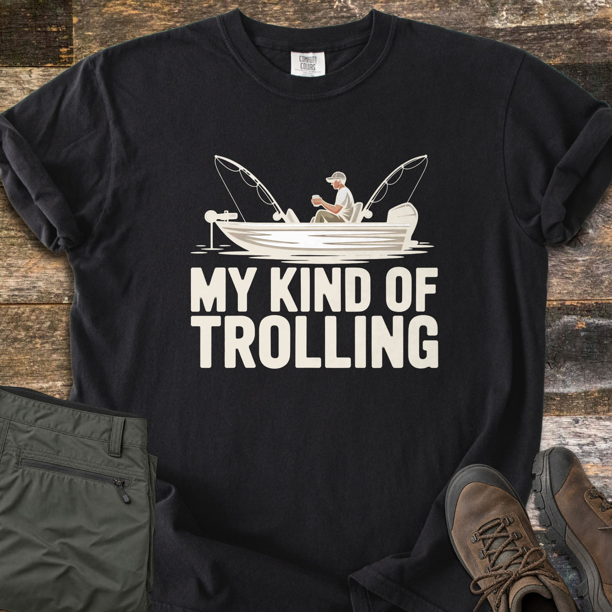 My Kind Of Trolling Comfort Colors T-shirt in Black, laid flat. A man in a cap holds a cup in a small fishing boat with two bent rods and motors, above the words My Kind Of Trolling in cream. Olive cargo pants and brown hiking boots sit beside it on weathered wood.