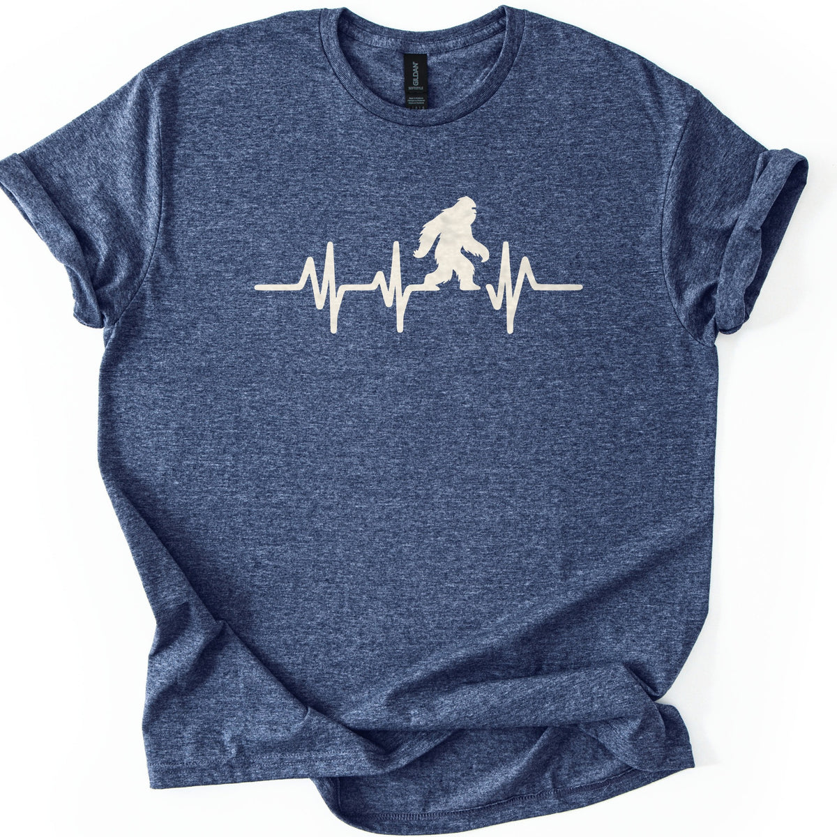 Yeti Heartbeat Comfort Colors T-shirt in Heather Navy, laid flat. A cream-colored heartbeat line runs across the chest, with a walking Bigfoot silhouette standing within it; there are no words. The sleeves are cuffed and the shirt lies on a white background.