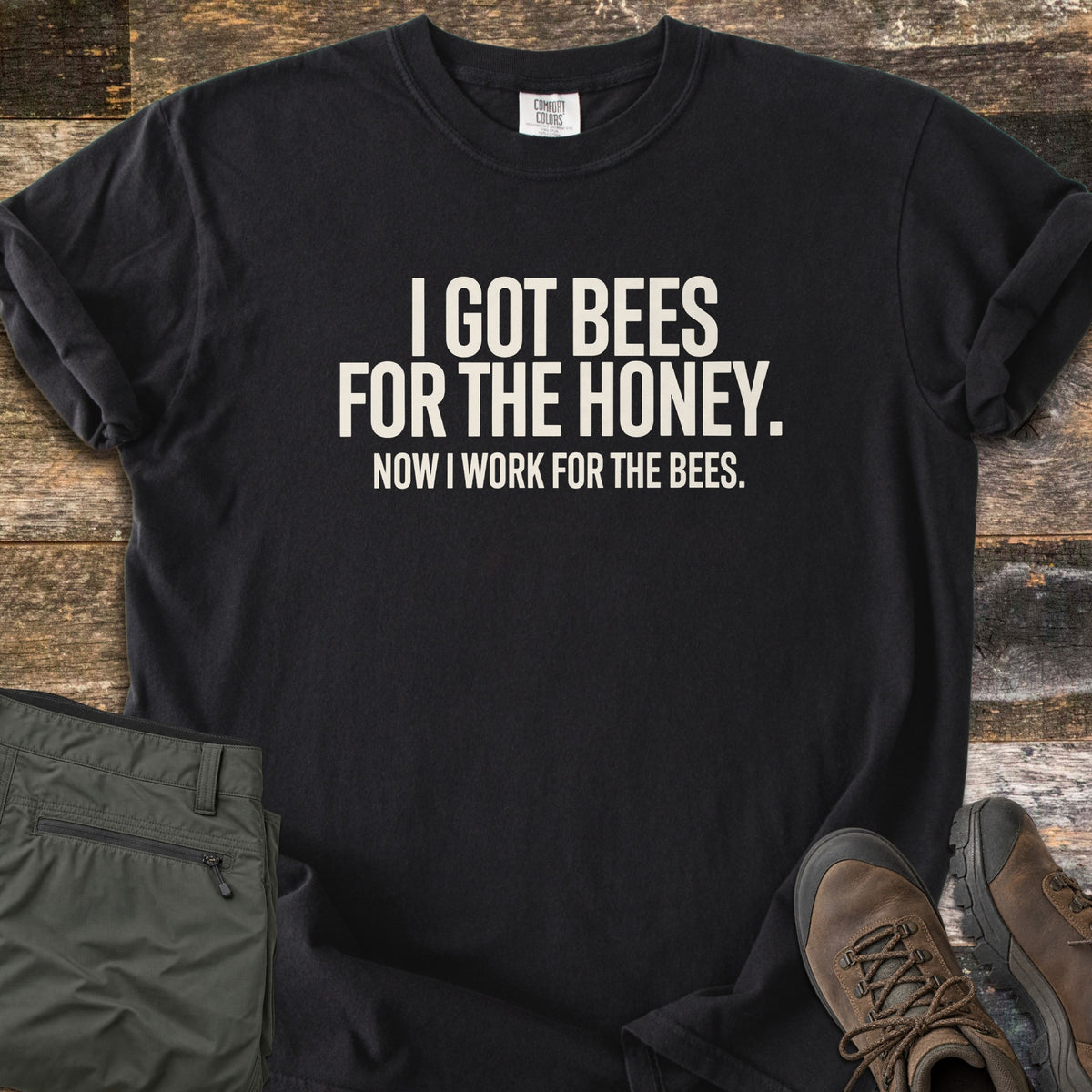 Work For The Bees Comfort Colors T-shirt in Black, laid flat. Cream block letters read I GOT BEES FOR THE HONEY. NOW I WORK FOR THE BEES. The shirt lies with cuffed sleeves on weathered wood planks beside folded olive green pants and brown hiking boots.