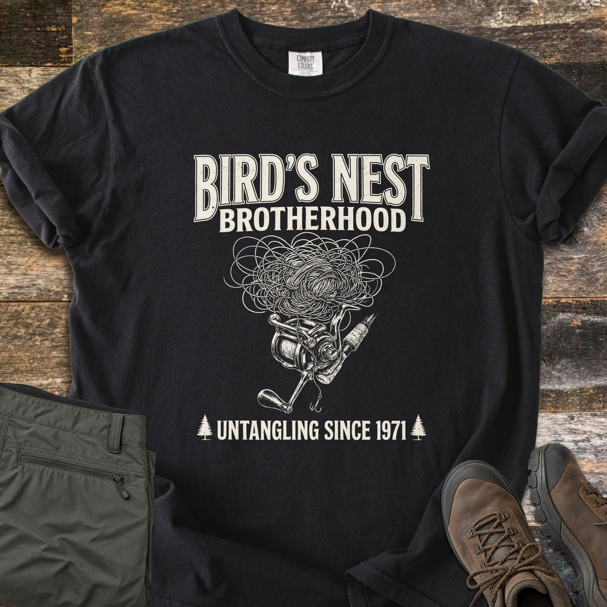Birds Nest Brotherhood Comfort Colors T-shirt in Black, laid flat. Cream sketch of a tangled line nest over a spinning reel, rod and hook, with Bird's Nest Brotherhood above and Untangling Since 1971 between pine trees below. Olive pants and hiking boots lie on old wood boards.