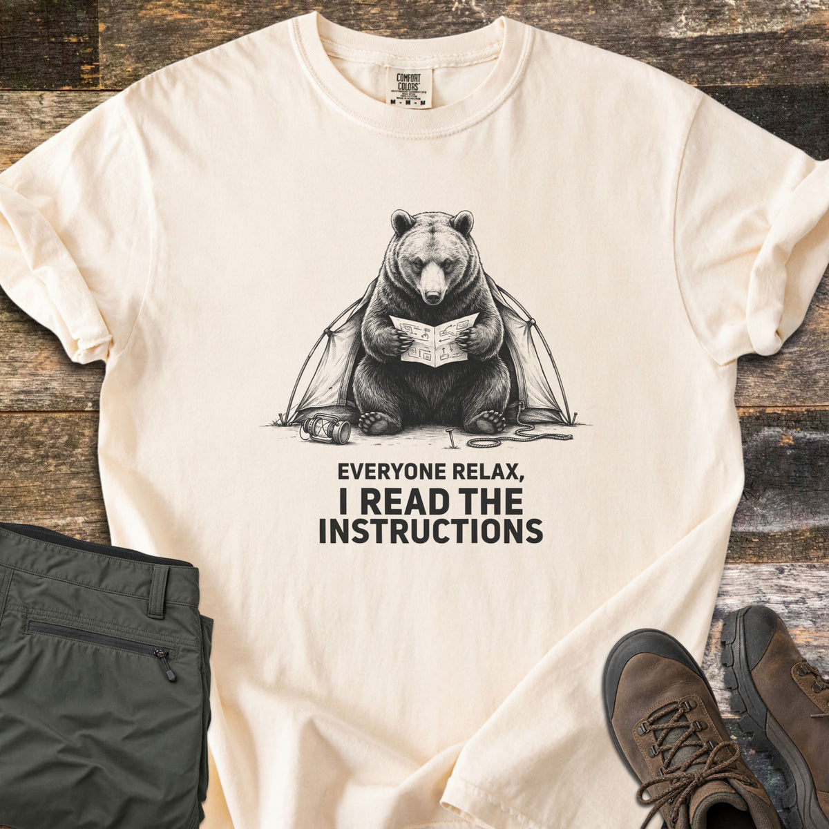 Everyone Relax I Read The Instructions Comfort Colors T-shirt in Ivory, laid flat. A black sketch of a bear reading instructions in front of a tent, with a tipped lantern and rope, above the words Everyone relax, I read the instructions. Olive cargo pants and brown hiking boots sit on dark wood.