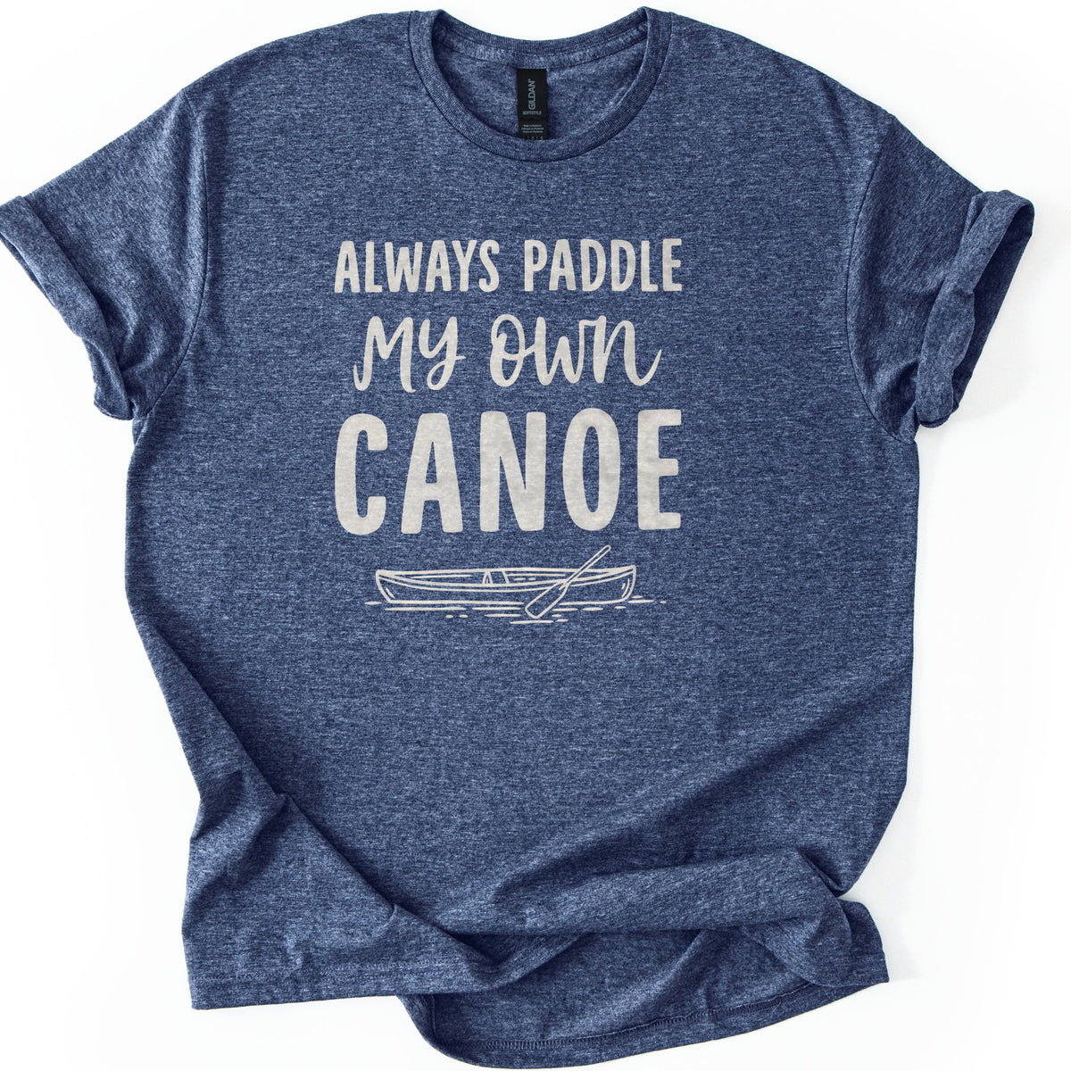 Paddle My Own Canoe Comfort Colors T-shirt in Heather Navy, laid flat. Cream text reads Always Paddle My Own Canoe above a line drawing of a canoe with a paddle on rippling water. The shirt has rolled sleeves and lies on a white surface.
