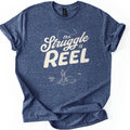 Struggle Is Reel Angler Comfort Colors T-shirt in Heather Navy, laid flat. Cream text reads the Struggle is REEL, with a fishing line arcing over the words to a hook. Below, a line drawing of an angler in a hat casts from a rock in the water. Sleeves cuffed, on a white background.