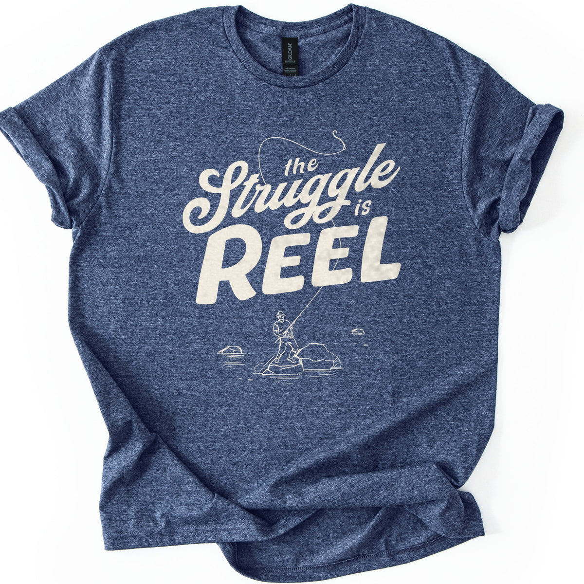 Struggle Is Reel Angler Comfort Colors T-shirt in Heather Navy, laid flat. Cream text reads the Struggle is REEL, with a fishing line arcing over the words to a hook. Below, a line drawing of an angler in a hat casts from a rock in the water. Sleeves cuffed, on a white background.