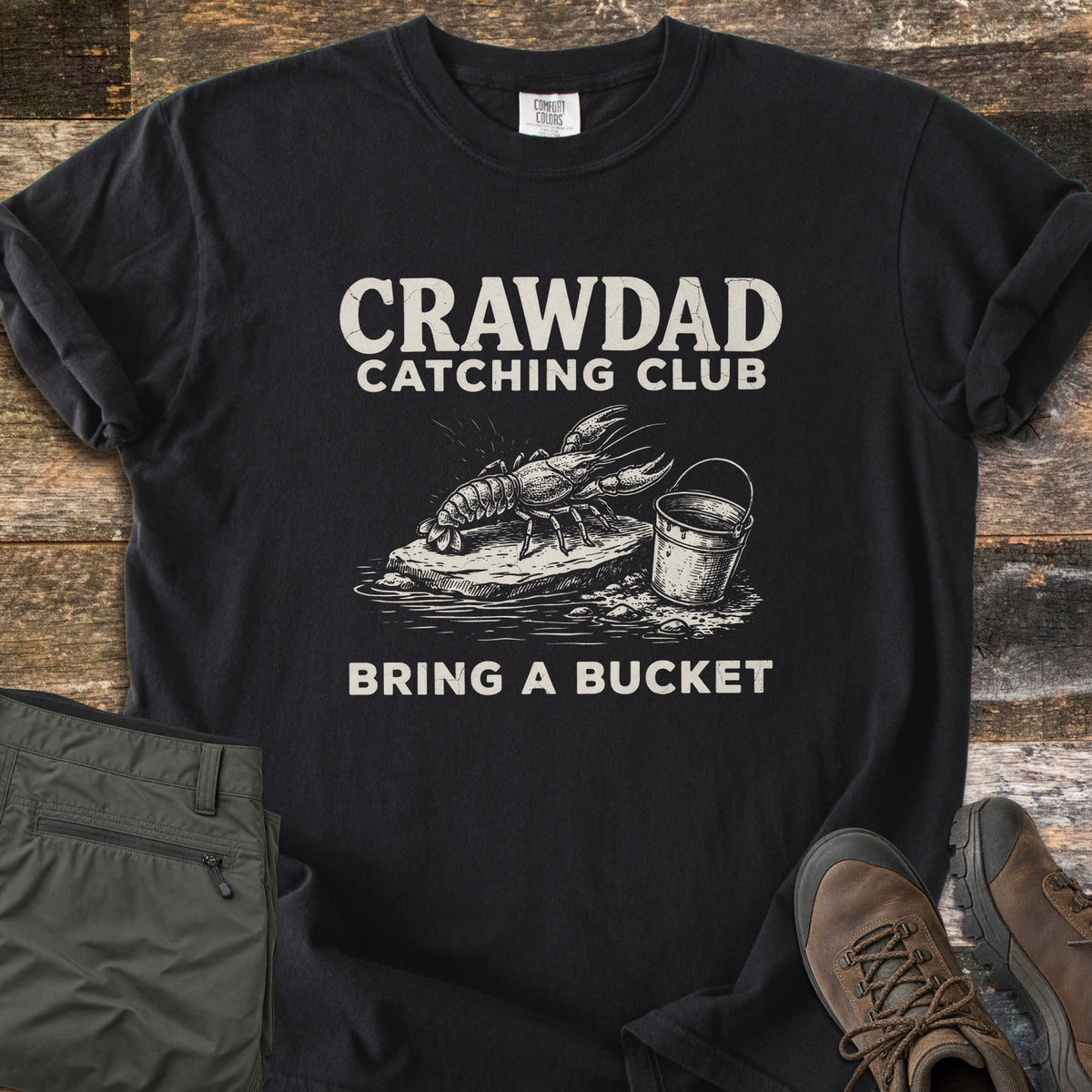Crawdad Catching Club Comfort Colors T-shirt in Black, laid flat. Cream print reads CRAWDAD CATCHING CLUB above a sketch of a crawdad on a rock beside a bucket, with BRING A BUCKET below. Folded olive pants and brown hiking boots lie beside it on weathered wood.