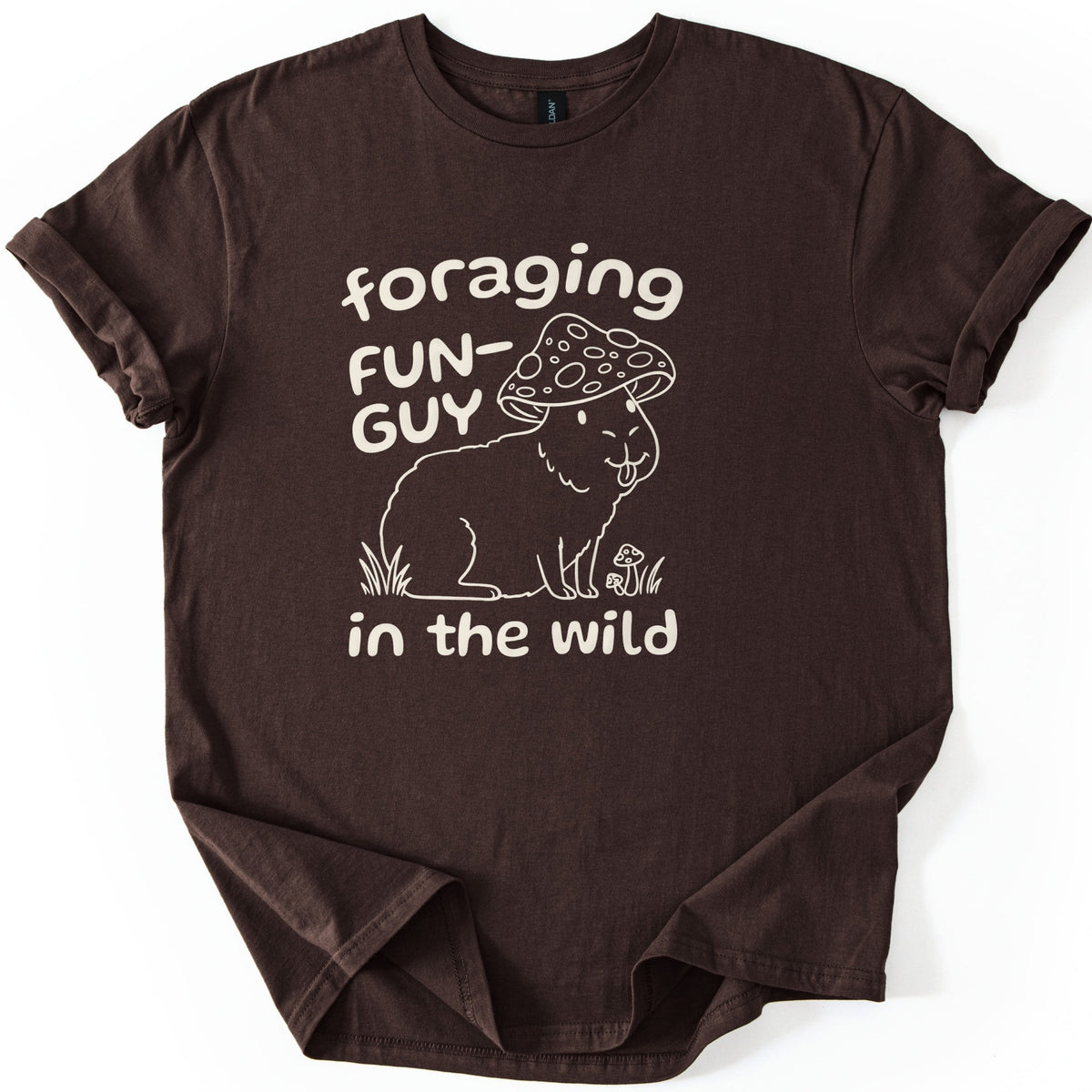 Foraging Fun-Guy Capybara Comfort Colors T-shirt in Dark Chocolate, laid flat. A cream line drawing of a winking capybara with its tongue out, wearing a spotted mushroom cap and sitting in grass by tiny mushrooms, with the words foraging FUN-GUY in the wild. Sleeves rolled, on white.