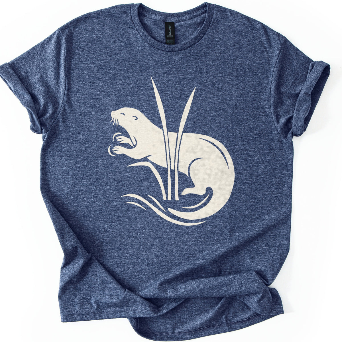 Otter In The Reeds Comfort Colors T-shirt in Heather Navy, laid flat. A cream silhouette of an otter with open mouth, whiskers and raised paws curls behind two tall reeds above swirling water lines. No words. The shirt lies on a white surface with sleeves rolled.