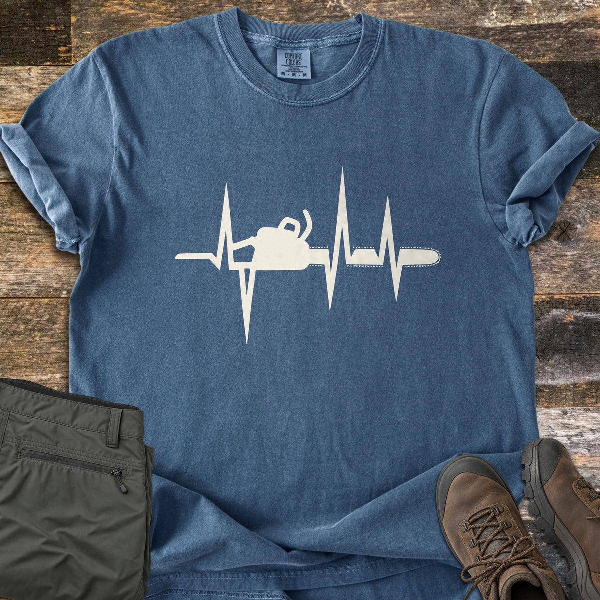 Chainsaw Heartbeat Comfort Colors T-shirt in Denim, laid flat. A cream heartbeat line runs across the chest with a chainsaw forming part of the pulse. Sleeves are cuffed on a weathered wood floor beside folded olive cargo pants and brown leather hiking boots.