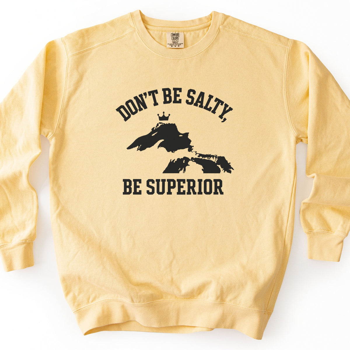 Be Superior Comfort Colors crewneck sweatshirt in Butter, laid flat. Black print of the words Don't Be Salty, arched above a silhouette of Lake Superior topped with a small crown, with Be Superior below. Laid on a white background.