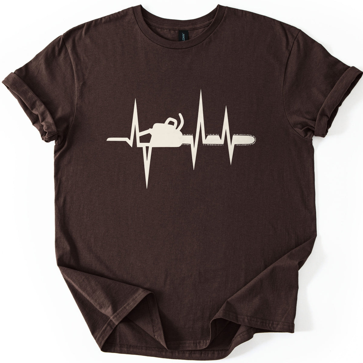Chainsaw Heartbeat Comfort Colors T-shirt in Dark Chocolate, laid flat. A cream heartbeat line with sharp peaks runs across the chest, with a chainsaw silhouette set into the line, its bar and chain extending to the right. The sleeves are rolled on a white background.