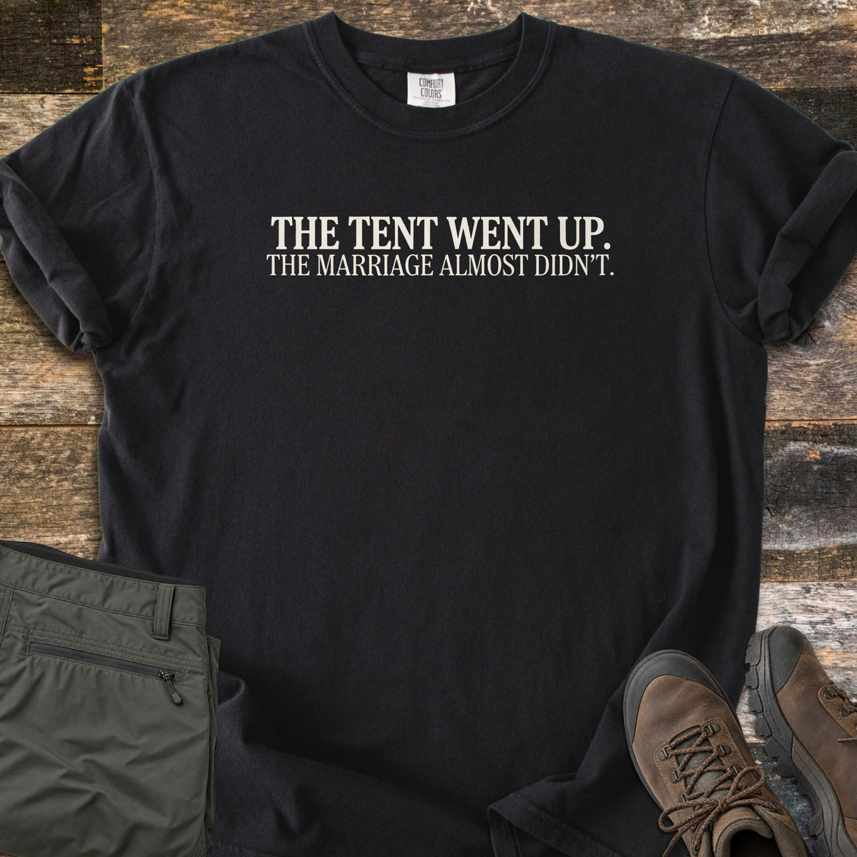 The Tent Went Up Comfort Colors T-shirt in Black, laid flat. Cream serif text across the chest reads The Tent Went Up. The Marriage Almost Didn't. Sleeves are rolled, with folded olive hiking pants and brown hiking boots beside it on weathered wood planks.