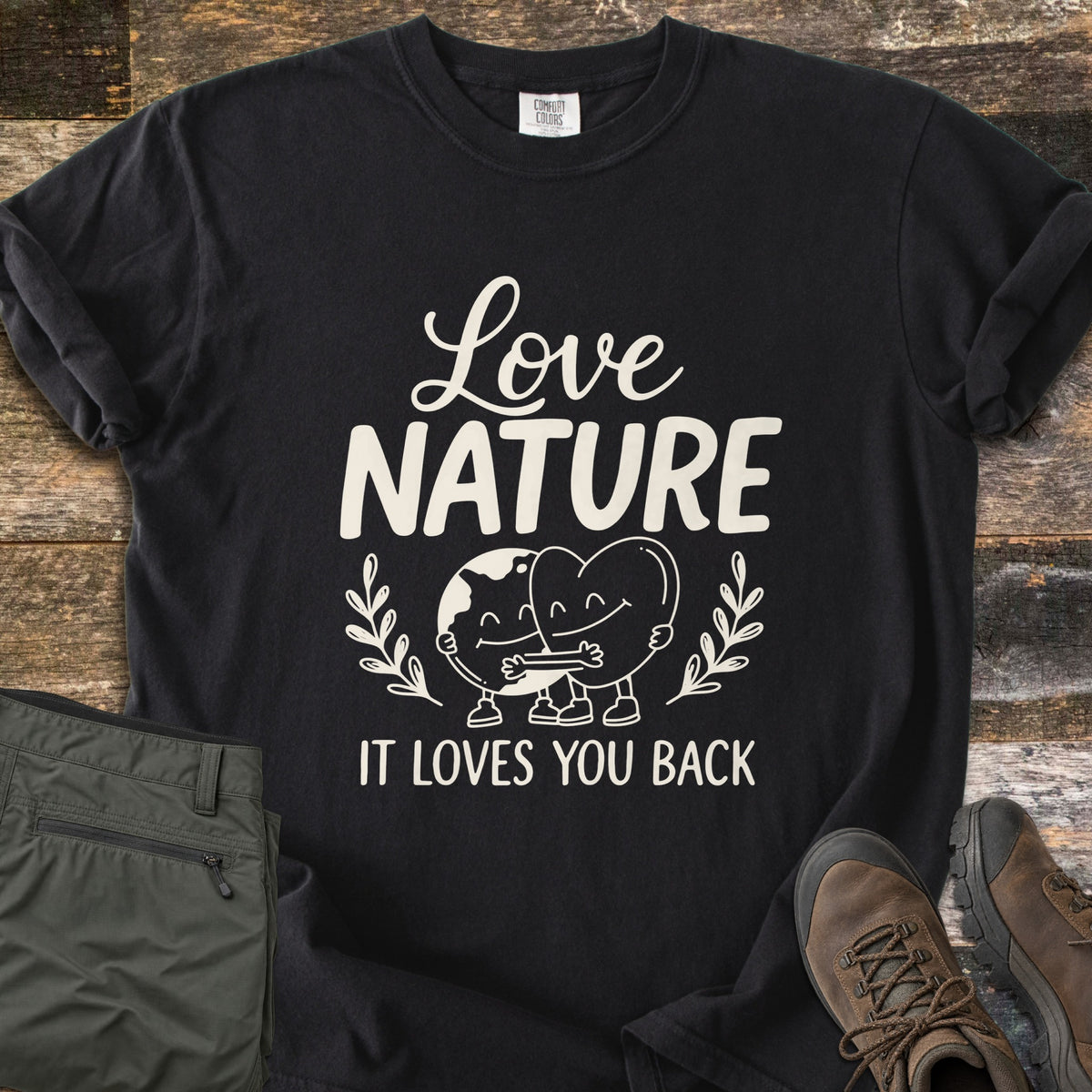 Nature Loves You Back Comfort Colors T-shirt in Black, laid flat. Cream print reads Love Nature above a smiling cartoon Earth and heart hugging between leafy sprigs, with It Loves You Back below. It lies on weathered wood beside olive hiking pants and brown boots.
