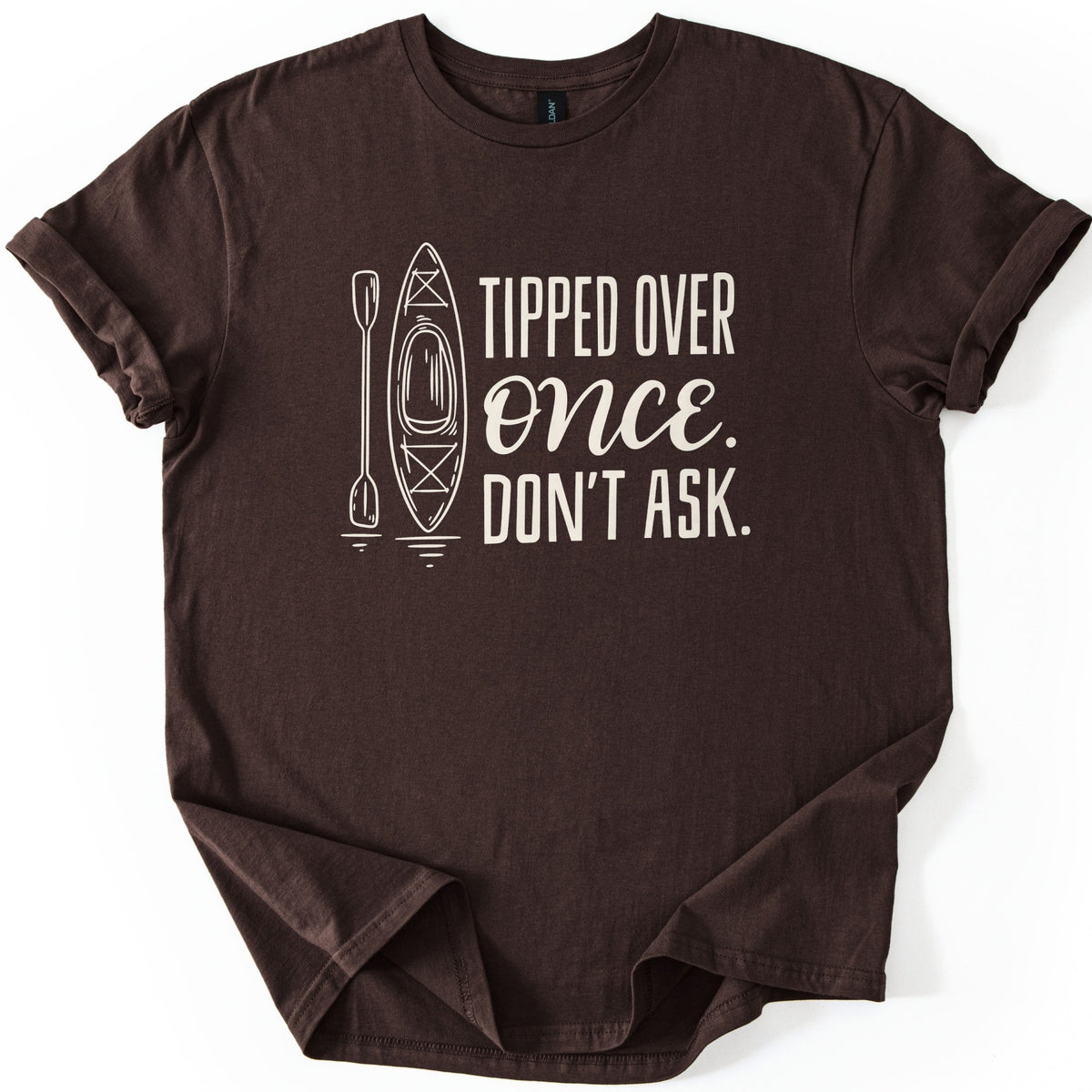 Tipped Over Once Kayak Comfort Colors T-shirt in Dark Chocolate, laid flat. A cream line drawing of an upright kayak and paddle with water ripples sits beside the words Tipped Over Once. Don't Ask., with Once in script. The shirt has cuffed sleeves on a white background.