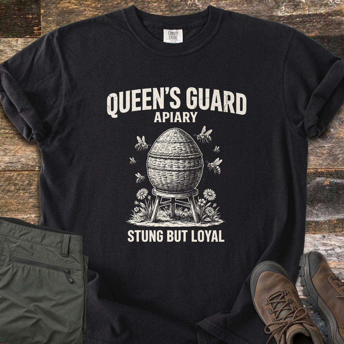 Queens Guard Apiary Comfort Colors T-shirt in Black, laid flat. A woven skep beehive on a wooden stand, circled by bees and wildflowers, sits under the words Queen's Guard Apiary and above Stung But Loyal in cream ink. Olive cargo pants and brown hiking boots lie on weathered wood.