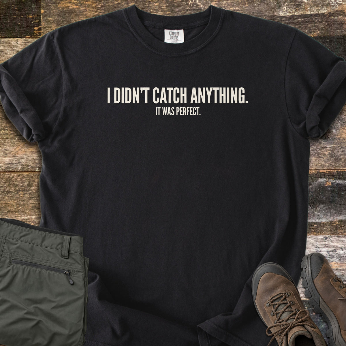 Didn't Catch Anything Comfort Colors T-shirt in Black, laid flat. Off-white block letters read I Didn't Catch Anything. with It Was Perfect. in smaller letters below. Sleeves are rolled, with folded olive hiking pants and brown hiking boots on weathered wood planks.