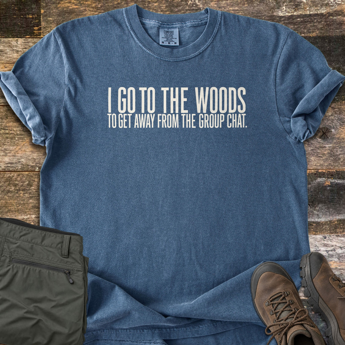 Away From The Group Chat Comfort Colors T-shirt in Denim, laid flat. White block text reads I GO TO THE WOODS, with TO GET AWAY FROM THE GROUP CHAT. below. The shirt lies on weathered wood planks beside olive hiking pants and brown lace-up hiking boots.