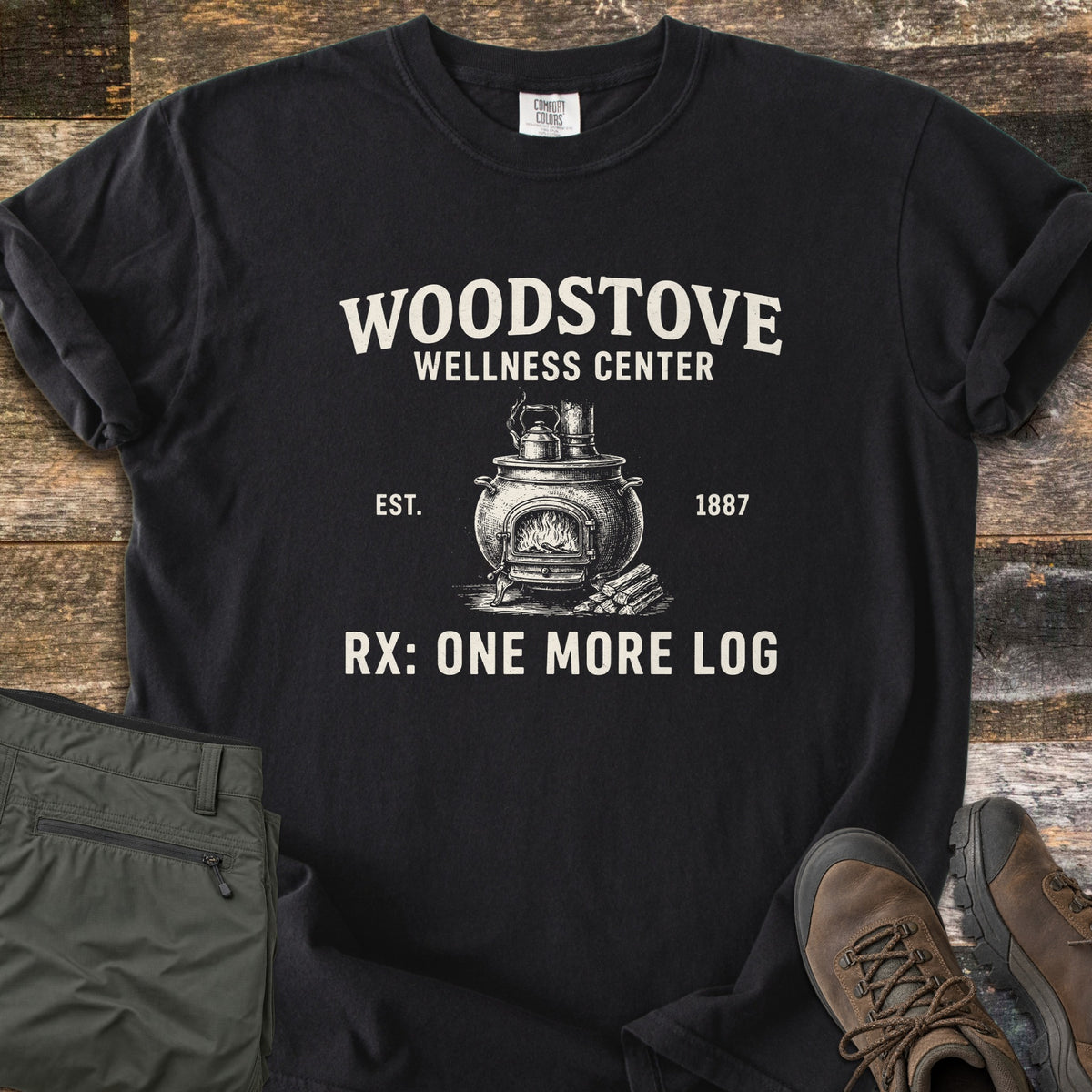 Woodstove Wellness Center Comfort Colors T-shirt in Black, laid flat. Cream print of a potbelly woodstove with a fire, a kettle on top and logs beside it, under Woodstove Wellness Center, flanked by Est. 1887, above Rx: One More Log. On wood planks with olive pants and brown hiking boots.