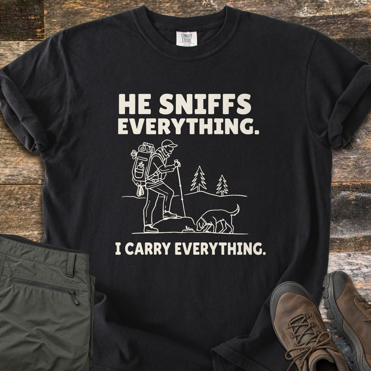 He Sniffs Everything. I Carry Everything Comfort Colors T-shirt in Black, laid flat. Cream line art of a hiker with a large backpack and trekking pole beside a dog sniffing a rock, between the words He Sniffs Everything. and I Carry Everything. Olive pants and brown hiking boots sit on weathered wood.