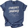 Embrace The Journey Trail Comfort Colors T-shirt in Heather Navy, laid flat. Cream hand-lettered words Embrace the Journey sit above a line drawing of two mountains, a round sun, and a winding road with tufts of grass. The heathered shirt has cuffed sleeves, on a white background.