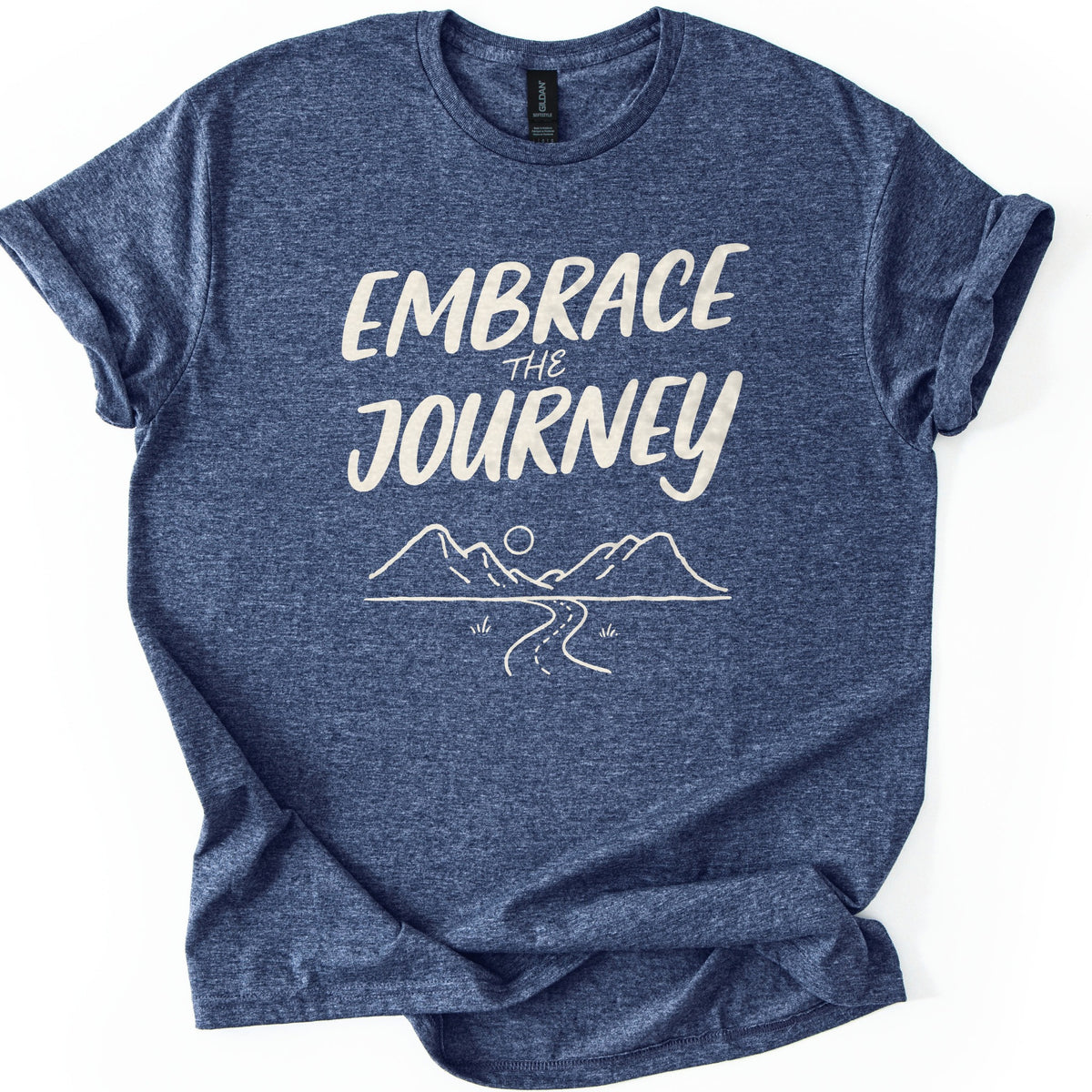 Embrace The Journey Trail Comfort Colors T-shirt in Heather Navy, laid flat. Cream hand-lettered words Embrace the Journey sit above a line drawing of two mountains, a round sun, and a winding road with tufts of grass. The heathered shirt has cuffed sleeves, on a white background.