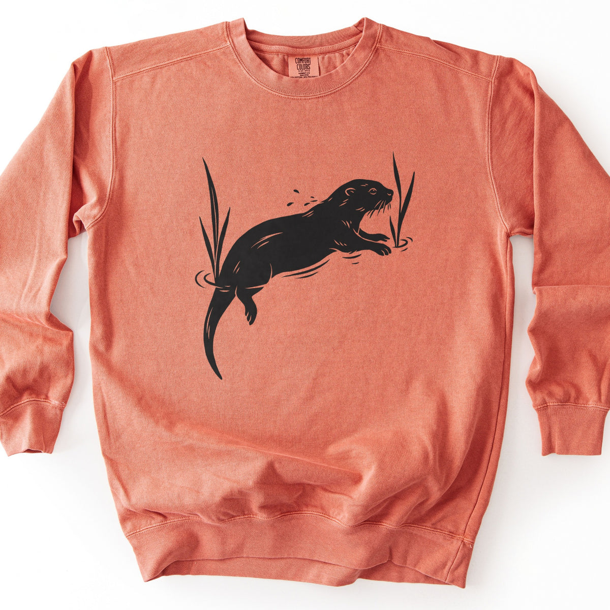 Swimming Otter Comfort Colors crewneck sweatshirt in Terracotta, laid flat. A black block-print style otter swims with its head raised, splashing droplets, between two tall reeds with ripples around them. No words are printed. The sweatshirt lies on a white background.