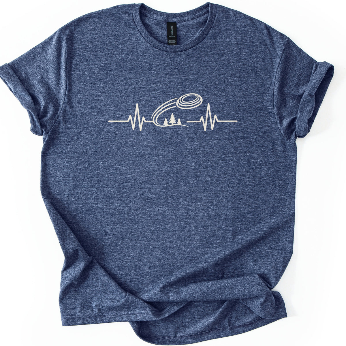 Trap Shooting Heartbeat Comfort Colors T-shirt in Heather Navy, laid flat. A cream heartbeat line with a clay target flying above three pine trees on a hill, with no words. The shirt lies on a white background with its sleeves rolled and the hem folded to one side.