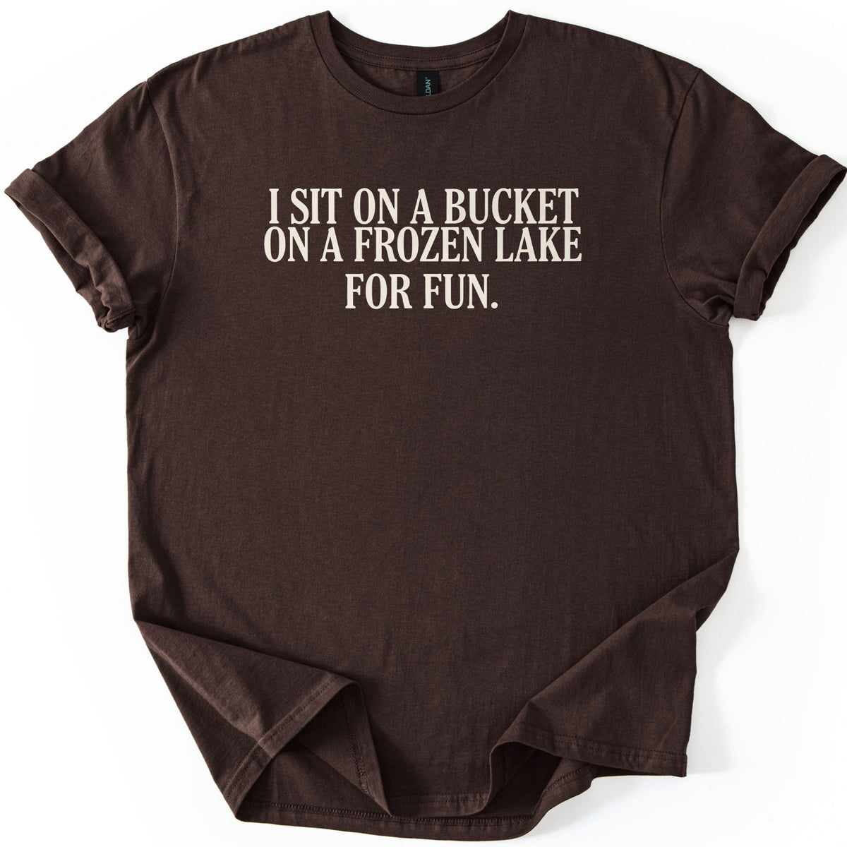 Bucket On A Frozen Lake Comfort Colors T-shirt in Dark Chocolate, laid flat. Cream serif capital letters across the chest read I SIT ON A BUCKET ON A FROZEN LAKE FOR FUN. The shirt lies on a white surface with its sleeves rolled and the hem loosely folded.