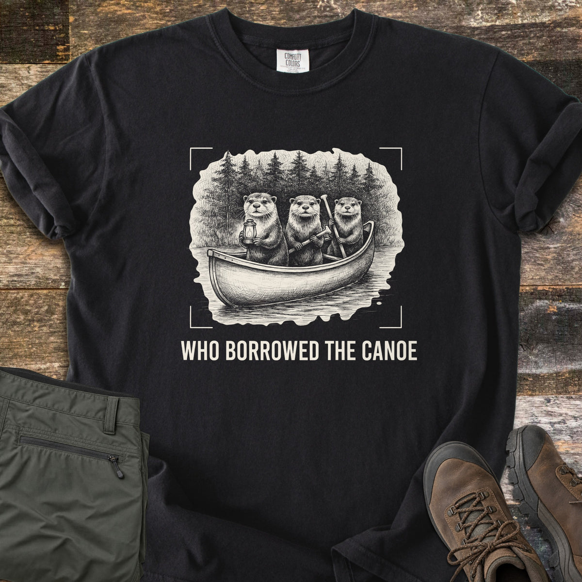 Who Borrowed The Canoe Comfort Colors T-shirt in Black, laid flat. A cream and black sketch of three otters in a canoe before pine trees, one holding a lantern, inside camera corner marks above the words Who Borrowed The Canoe in cream. Olive pants and brown boots sit on wood.