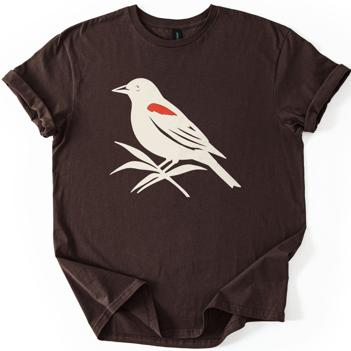 Red Winged Blackbird Comfort Colors T-shirt in Dark Chocolate, laid flat. A cream silhouette of a red-winged blackbird with a red shoulder patch, perched on slender leaves, with no words. The shirt has rolled sleeves and lies on a white background.