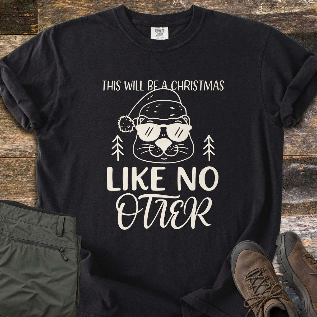 Christmas Like No Otter Comfort Colors T-shirt in Black, laid flat. A cartoon otter in a Santa hat and sunglasses sits between two pine trees, with This Will Be a Christmas above and Like No Otter below, in cream. Olive cargo pants and brown hiking boots lie on weathered wood.