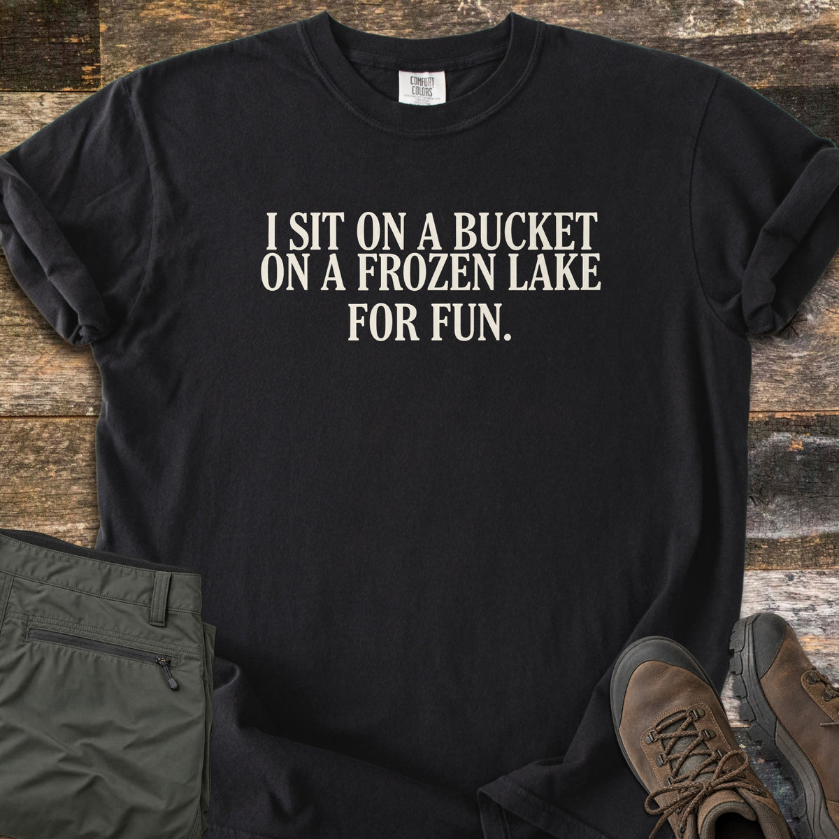 Bucket On A Frozen Lake Comfort Colors T-shirt in Black, laid flat. Cream serif text reads I SIT ON A BUCKET ON A FROZEN LAKE FOR FUN. The shirt has rolled sleeves and lies on weathered wood planks beside folded olive hiking pants and brown hiking boots.