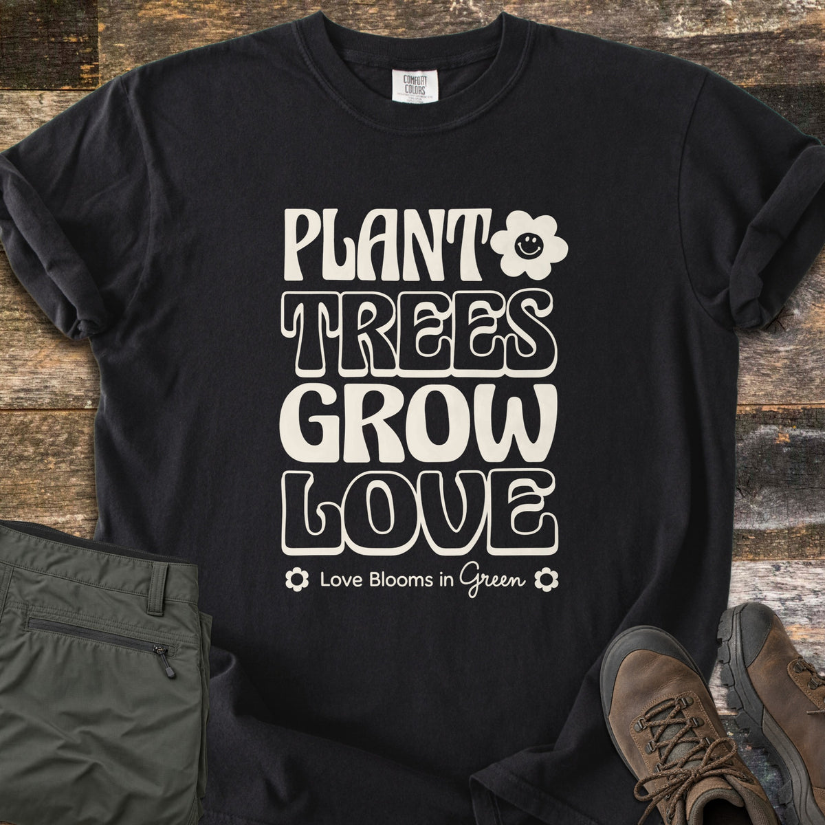 Plant Trees Grow Love Comfort Colors T-shirt in Black, laid flat. Cream retro bubble letters read Plant Trees Grow Love beside a smiling daisy, above Love Blooms in Green. Sleeves cuffed on weathered wood with folded olive hiking pants and brown hiking boots.