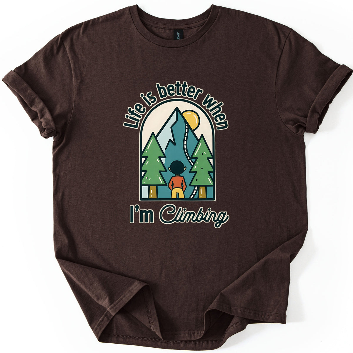 Better When I'm Climbing Comfort Colors T-shirt in Dark Chocolate, laid flat. The words Life is better when arch over a window scene of a person facing a teal mountain with a winding trail, a yellow sun and two pine trees, with I'm Climbing below. Sleeves rolled, on a white surface.