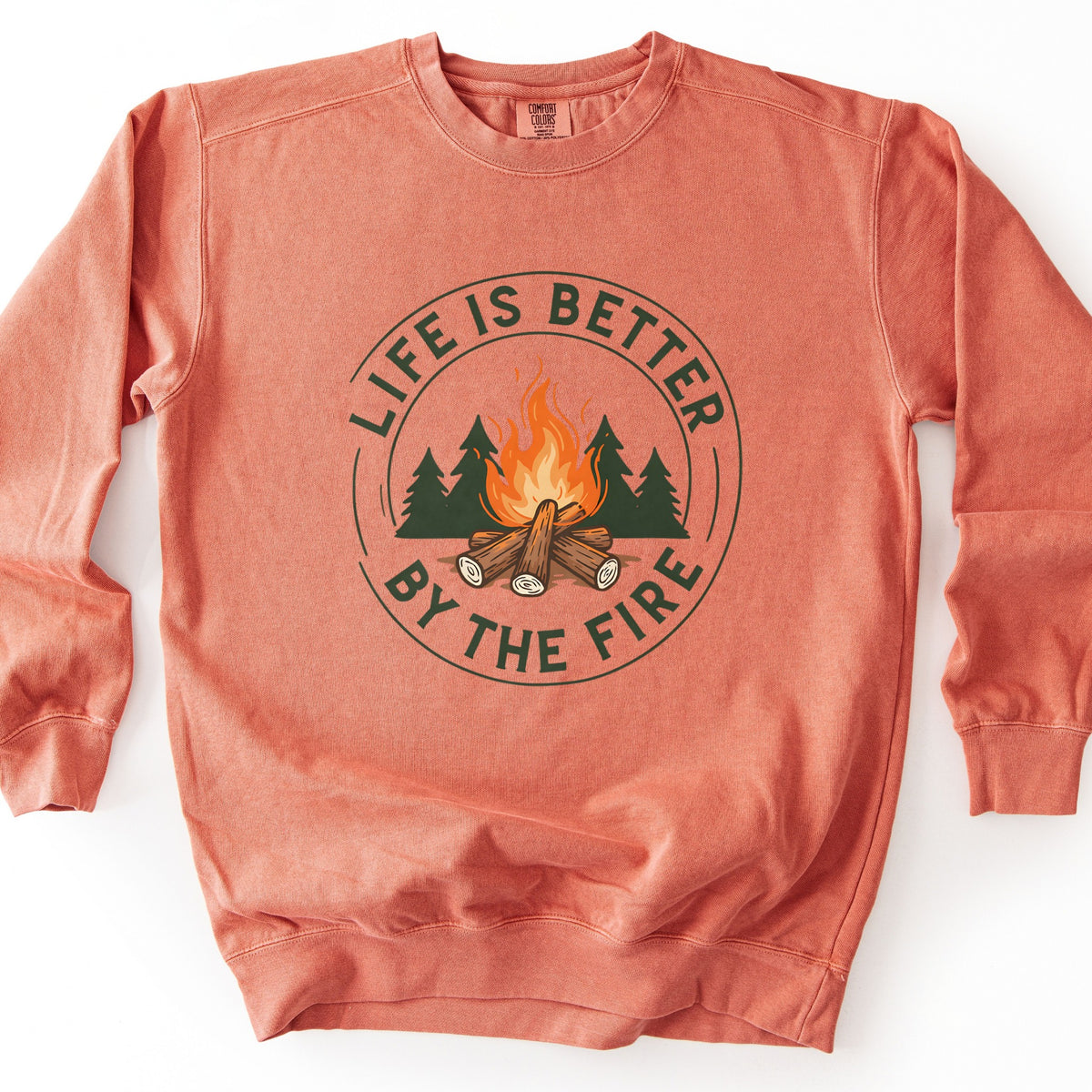 Life Is Better By The Fire Ring Comfort Colors crewneck sweatshirt in Terracotta, laid flat. A campfire of stacked logs with orange flames sits in front of dark green pines inside a dark green ring, with the words Life Is Better By The Fire curved around it. It lies on a white surface.