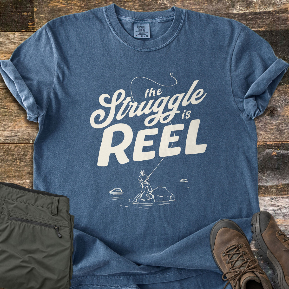 The Struggle Is Reel Comfort Colors T-shirt in Denim, laid flat. Cream text reads the Struggle is REEL under an arcing fishing line with a hook, above a sketch of an angler casting from a rock in water. Folded olive pants and brown hiking boots sit on wood planks.