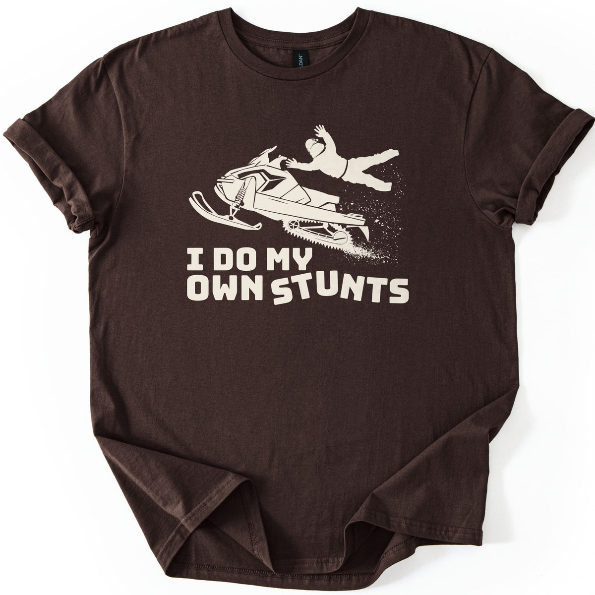 Snow Mobile Stunts Comfort Colors T-shirt in Dark Chocolate, laid flat. A cream silhouette of a snowmobile flying through the air, its rider thrown off behind it in a spray of snow, above the words I Do My Own Stunts. The shirt lies on a white background with rolled sleeves.